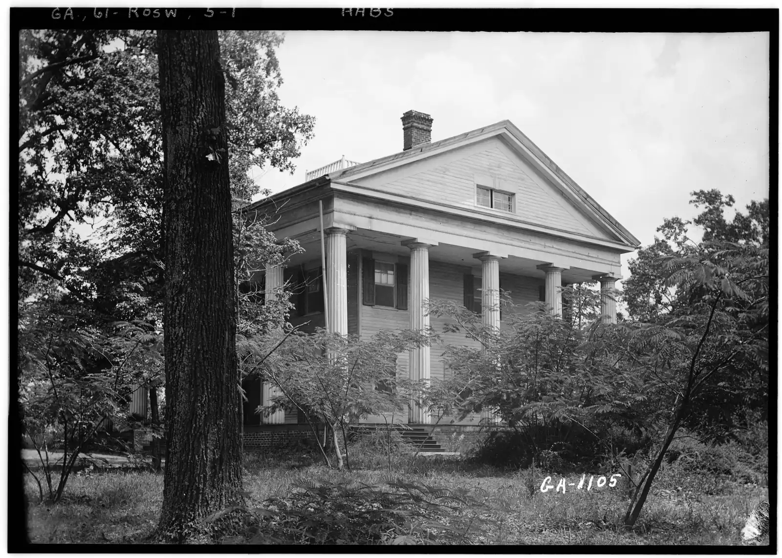 A black-and-white archival photograph of a white temple-form Greek Revival house with fluted Doric columns, a deep pediment with an attic window, a brick chimney and a railed rooftop walk, seen at an angle behind a large dark oak trunk on the left and feathery mimosa-like shrubs and long grass in the foreground. The film edge is lettered "GA, 61-ROSW, 5-1" and "HABS", with "GA-1105" written in the lower right.