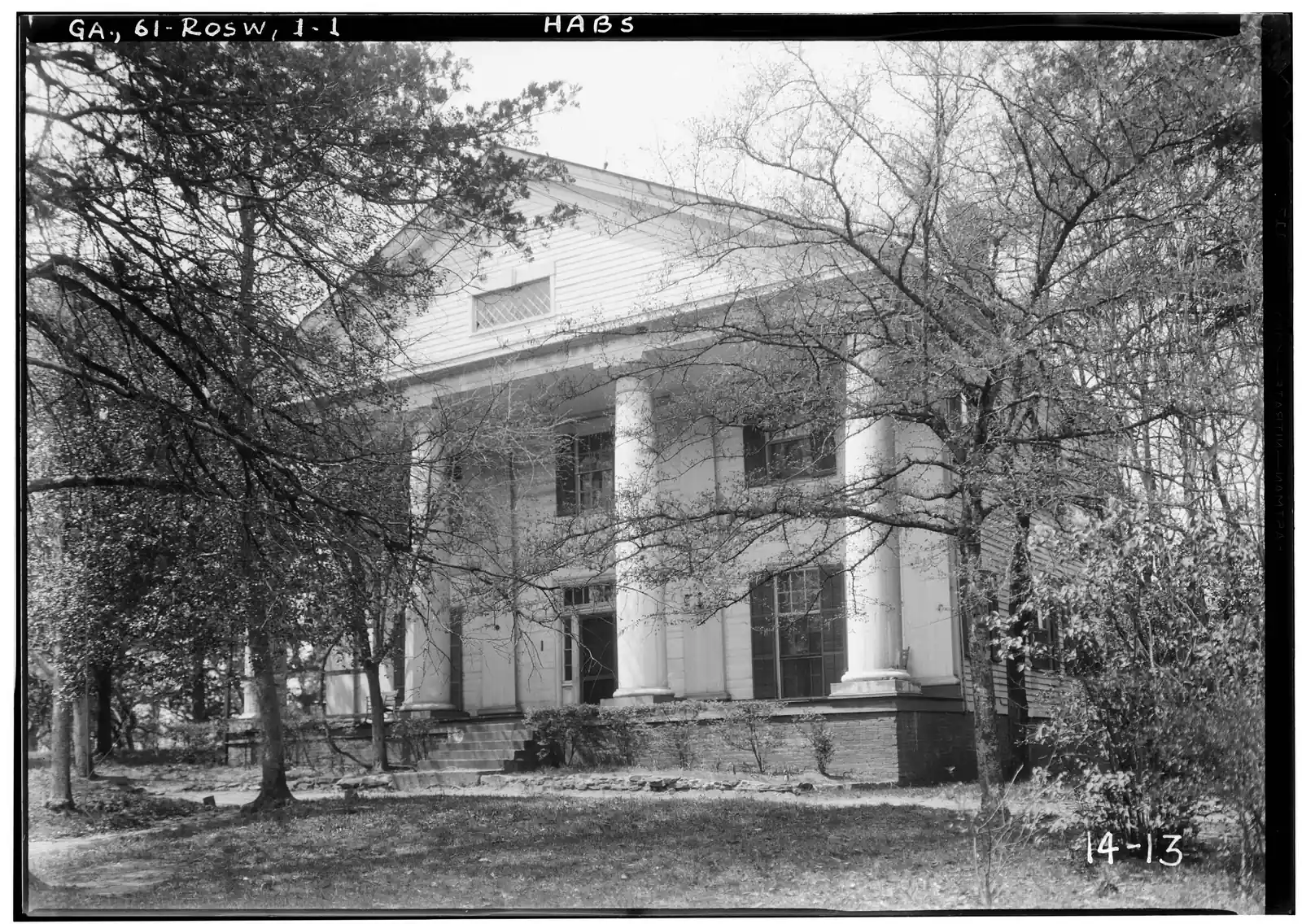 A black-and-white archival photograph of a white Greek Revival house with a pedimented front portico of thick round columns, seen at an angle through a lattice of bare and budding tree branches. It has a diamond-pane attic window in the pediment, tall shuttered windows and a brick foundation with front steps. The film edge is lettered "GA, 61-ROSW, 1-1" and "HABS", with "14-13" written at lower right.