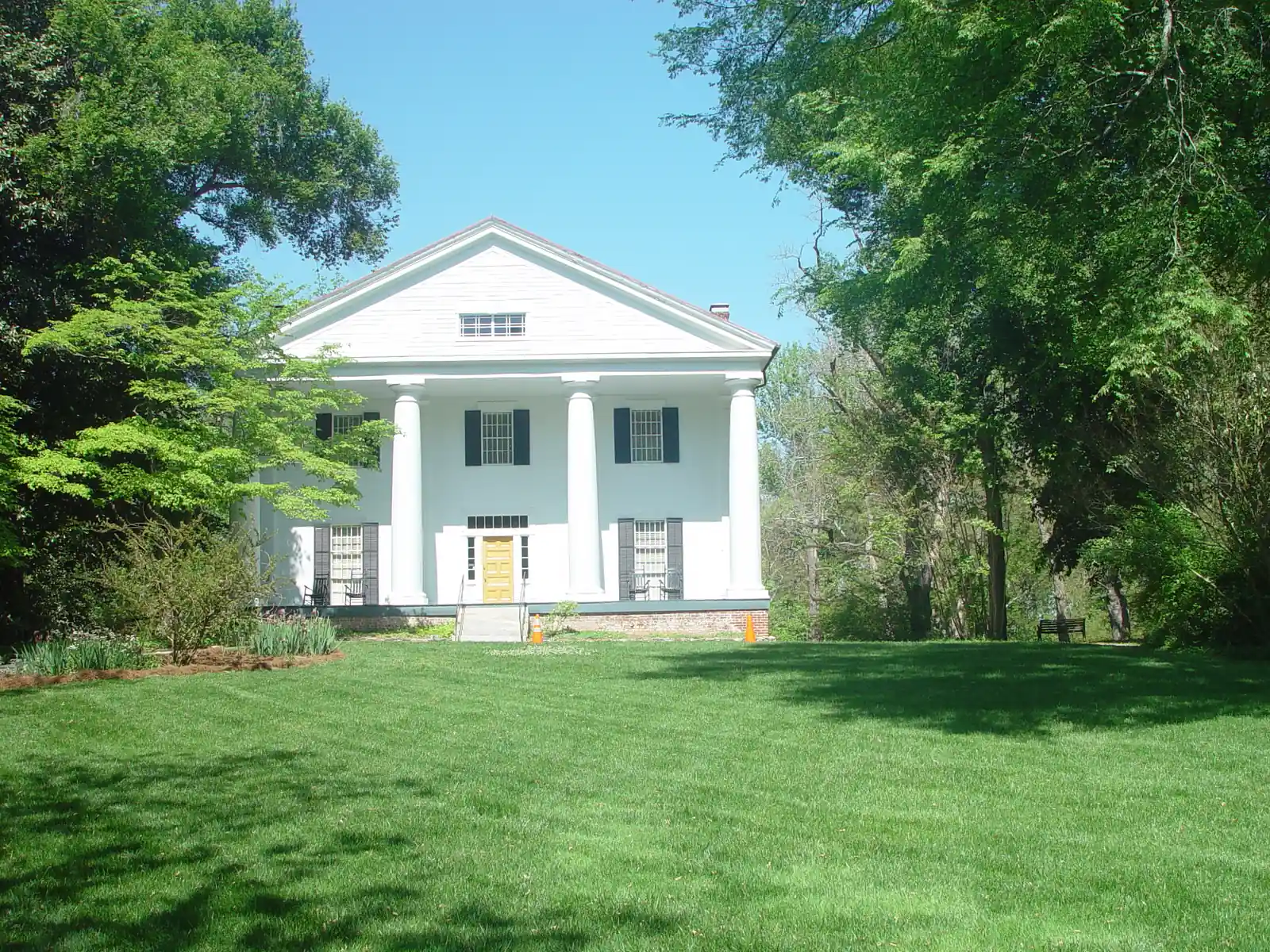 A white two-storey Greek Revival house with a full-height front portico of four thick round columns under a broad triangular pediment with a small attic window, black shutters, a mustard-yellow front door and two rocking chairs on each side of the porch, on a brick foundation. It sits at the top of a wide, freshly mown green lawn framed by large spring-green trees, under a clear blue sky. Two orange traffic cones flank the front steps and a bench stands at the right.