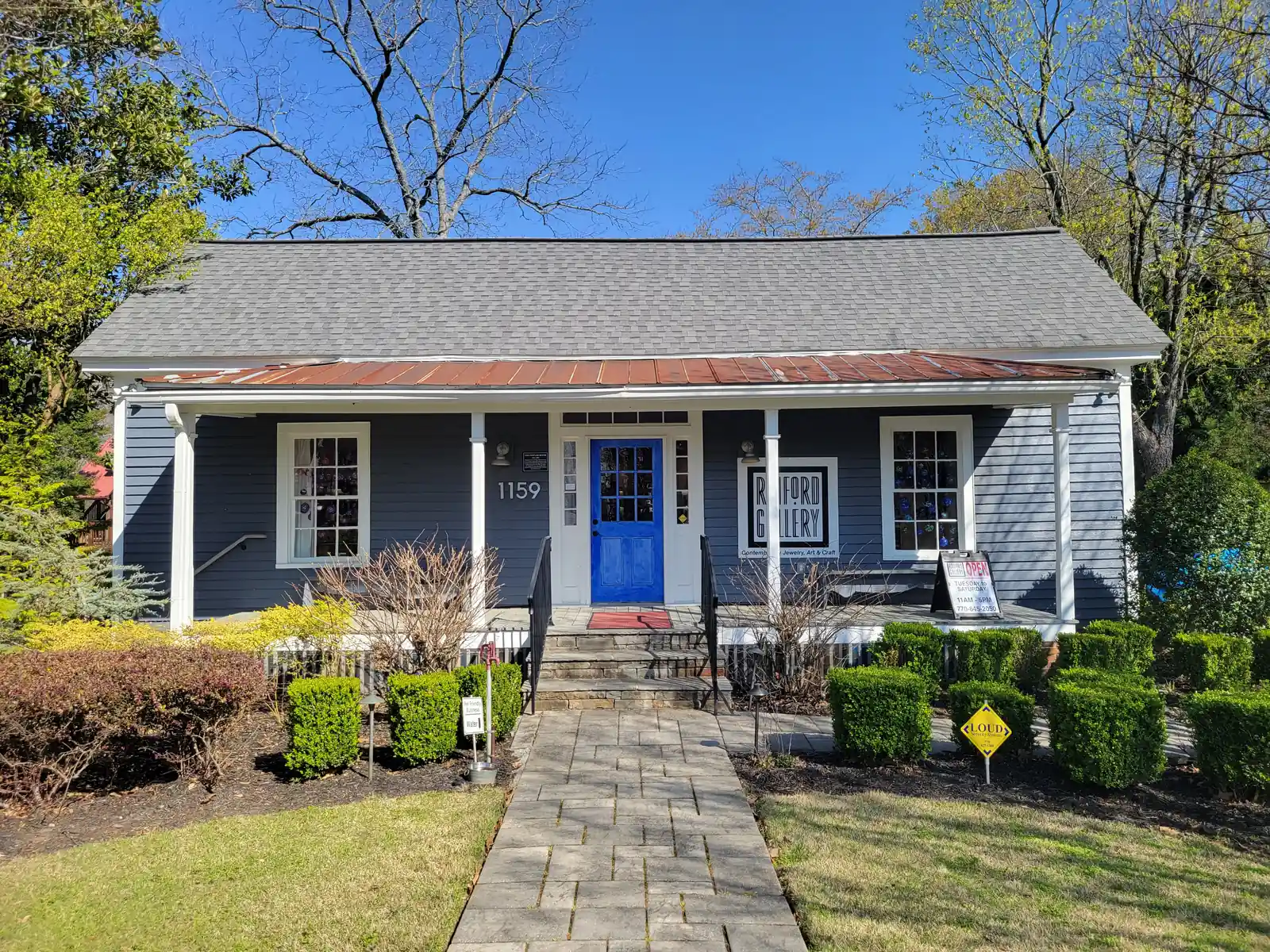 Symmetrical front view of a one-storey slate-blue clapboard cottage with white trim, a rust-red metal porch roof, a bright cobalt-blue front door and house number 1159. A stone path leads up through clipped boxwoods to the porch. A sign in the right window reads "Raiford Gallery, Contemporary Jewelry, Art & Craft" with an A-frame "Open Tuesday to Saturday" board beside it. A small black plaque by the door is titled "The Fowler House". Bare and budding trees stand against a clear blue sky.