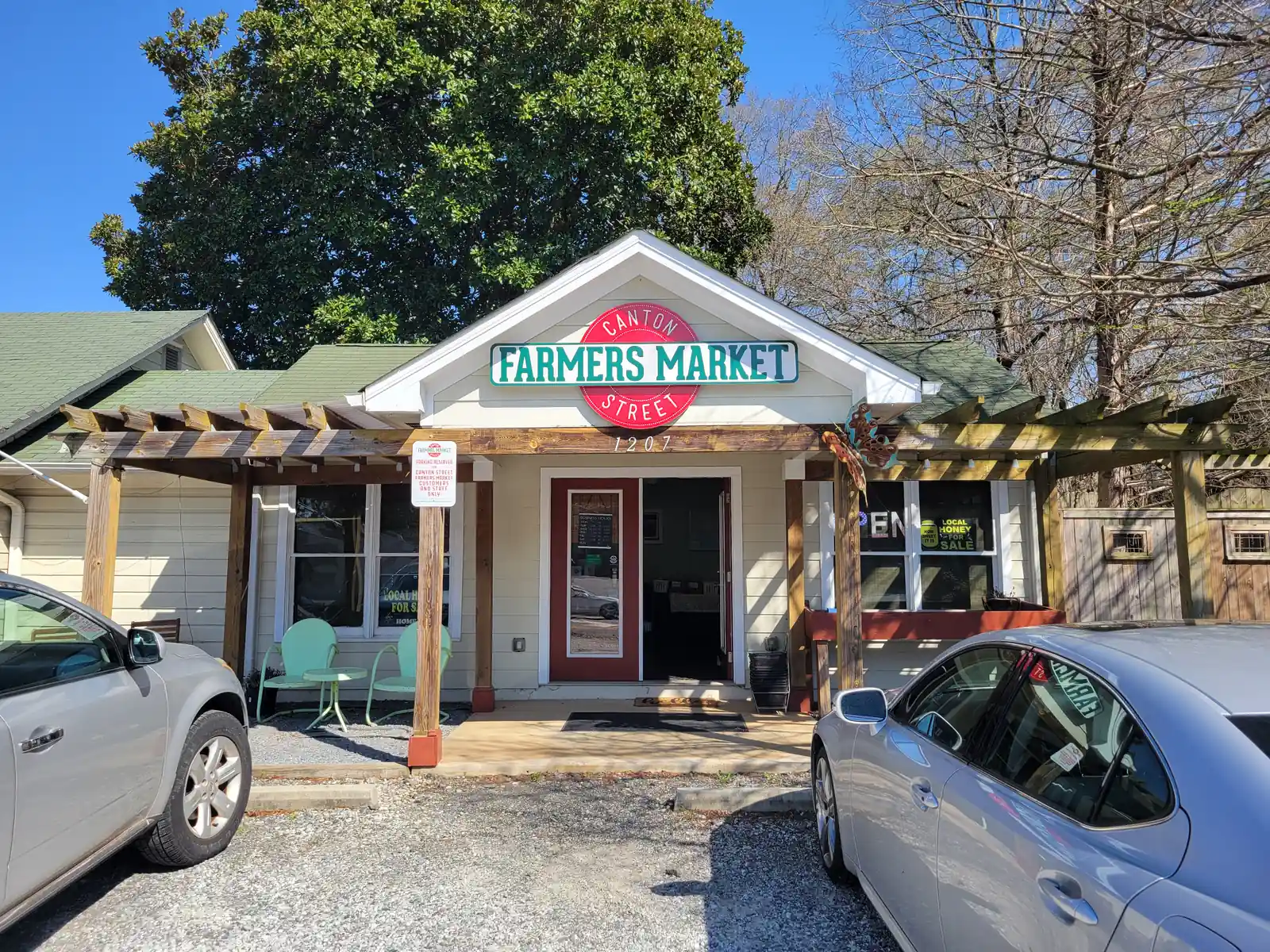 Front of a small cream clapboard cottage with a gabled entry and a wooden pergola. A large sign reads "Canton Street Farmers Market" with house number 1207 below it. The front door is open, and there are two mint-green metal chairs and a little table on the porch, an "OPEN" sign and a "Local Honey for Sale" sign in the windows, and a "Parking reserved for Canton Street Farmers Market customers" sign on a post. Two silver cars are parked on the gravel in front. A big magnolia and bare trees stand behind under a clear blue sky.