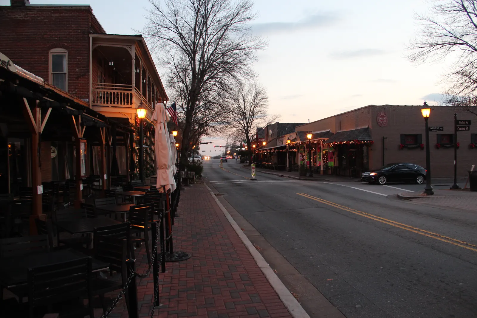 Dusk view along a brick-paved sidewalk lined with stacked black café chairs, tables and closed white patio umbrellas under a timber-framed awning; above it rises a two-storey red-brick building with a white Victorian-style upper gallery, glowing iron lamp posts and an American flag. Across the street a low row of brick storefronts is strung with holiday lights and red bows, a black sedan turns at the corner, and a street sign on the right reads "Webb St" / "Canton St" with a "Webb St Shops" arrow. Bare winter trees, pale pink sky.