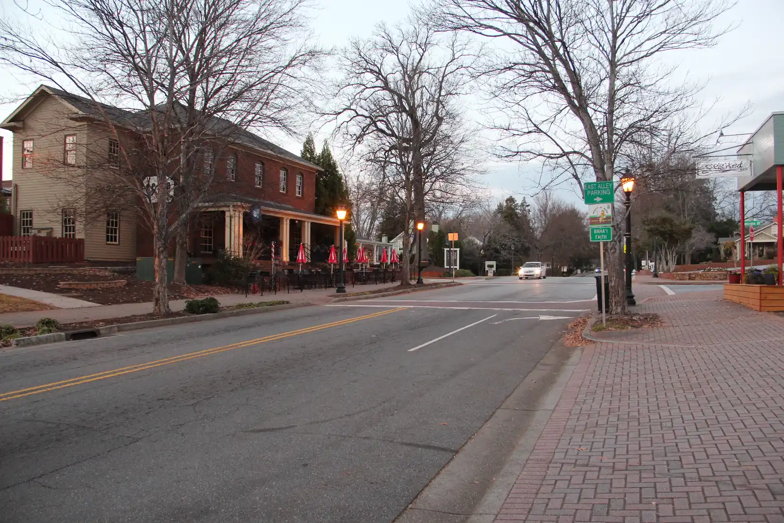 Early-evening street scene: an empty two-lane street with double yellow lines and a brick-paved sidewalk on the right. On the left stands a two-storey red-brick building with arched upper windows and a columned front porch, with a patio of black tables and closed red umbrellas; a tan clapboard building is beside it. On the right are an iron lamp post, a green "East Alley Parking" sign, an Adopt-a-Road sign and a "Baha'i Faith" direction sign, and a hanging shop sign reading "Spirited" under a red-posted porch. One car with its headlights on approaches, and bare trees stand against an overcast sky.
