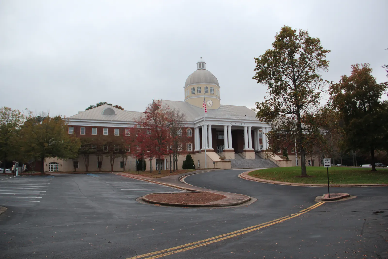 A large red-brick civic building with cream trim, a central white columned portico with "CITY HALL" lettered on the entablature, a flagpole with an American flag, and a white drum and dome with a clock and cupola, seen across an empty curving asphalt drive and parking lot. Autumn-tinted trees and a small "Keep Right" sign are in the middle distance. Overcast sky.