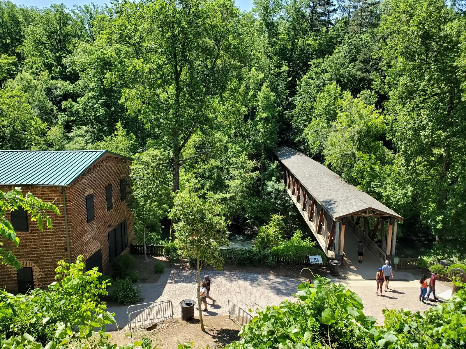 High overhead view in bright late-spring sun: on the left, a three-storey red-brick mill building with a green standing-seam metal roof and dark shutters; at centre right, a long timber covered footbridge with a grey shingle roof and diagonal truss sides runs from a paved brick plaza into dense green forest across a rushing creek, with white water visible below. About eight visitors, small and unidentifiable, walk on the plaza and enter the bridge. An interpretive panel and a trash can stand by the bridge entrance.