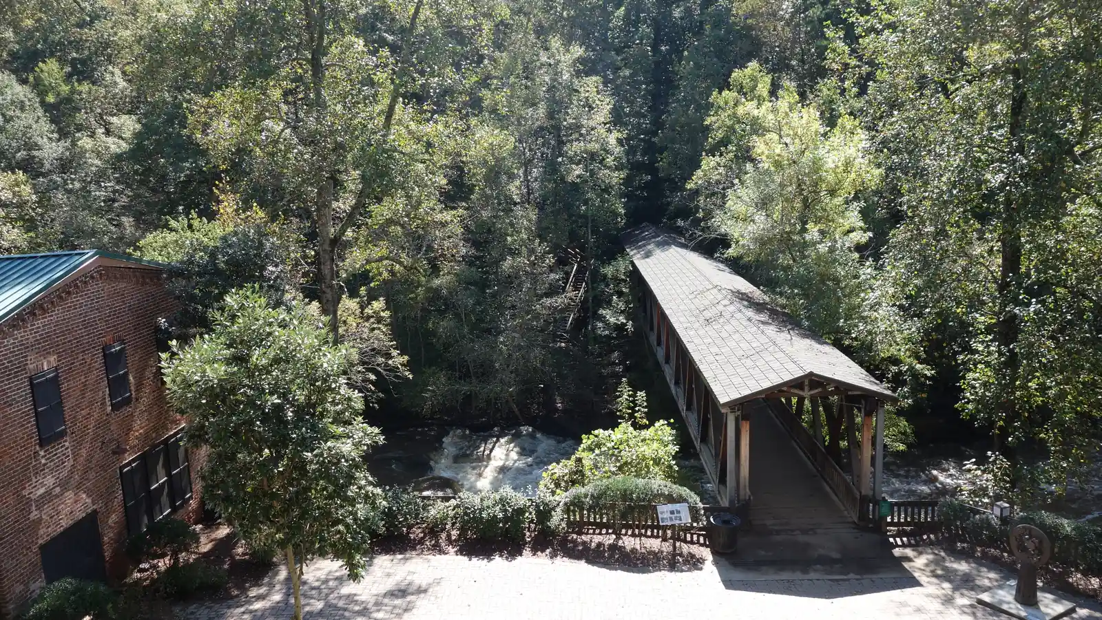 Elevated view in dappled autumn light: on the left, the red-brick wall of a mill building with a green metal roof and black shutters; at centre right, a covered timber footbridge with a shingled roof and diagonal trusses crosses a creek where white water churns over dark rock ledges. A wooden stairway climbs the wooded far bank. In the foreground are a brick-paved plaza, an interpretive panel on a post, a trash can, and at the lower right corner a small sculpture made of a spoked iron wheel on a stone plinth. No people.