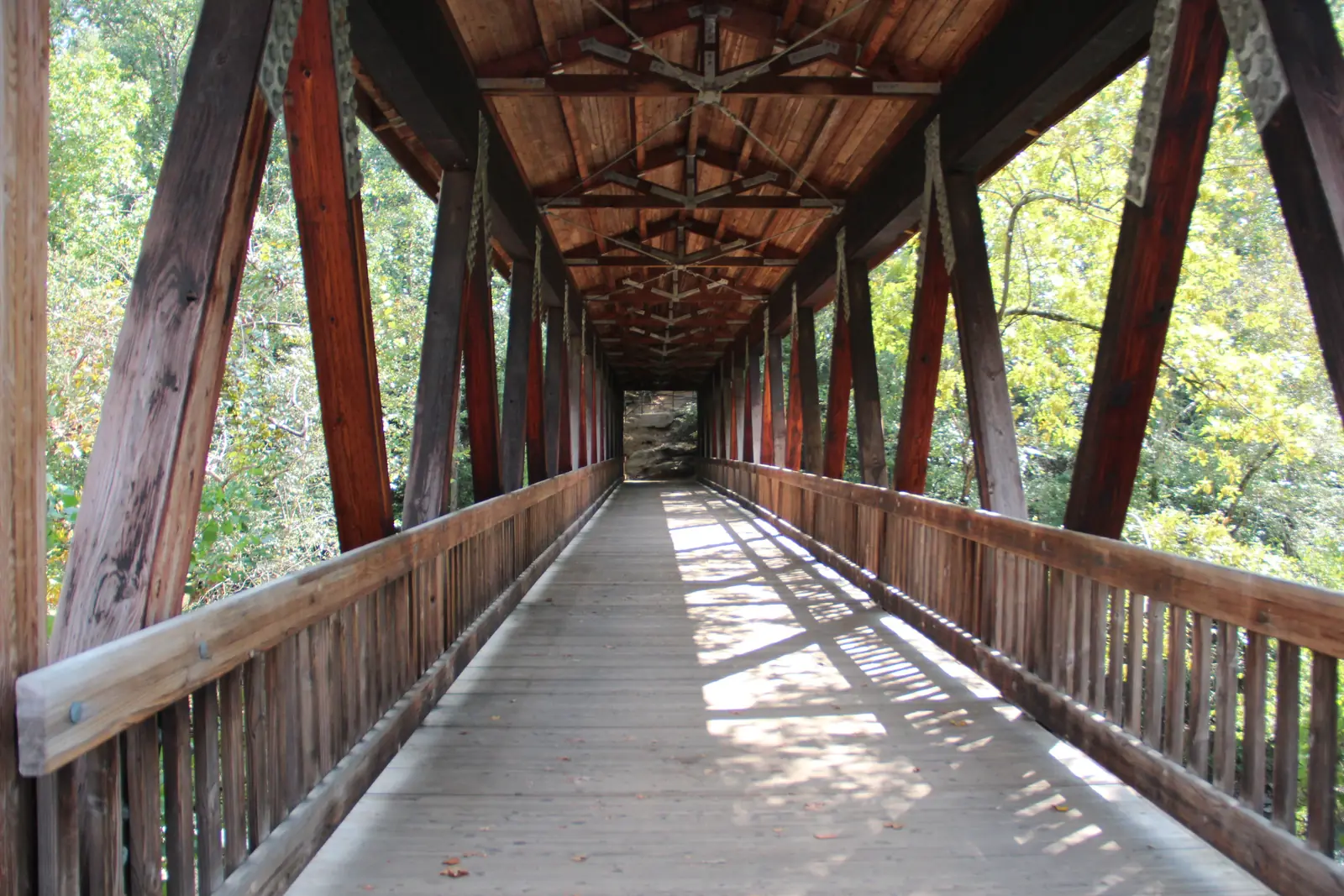 Symmetrical one-point-perspective view straight down the inside of a long timber covered footbridge: reddish diagonal timber trusses and galvanised steel plates on both sides, an exposed wooden roof structure overhead, slatted wooden railings and a plank deck striped with sun and shadow, leading to a small bright opening at the far end with rock and trees beyond. Green and yellow foliage shows through the open sides. No people.