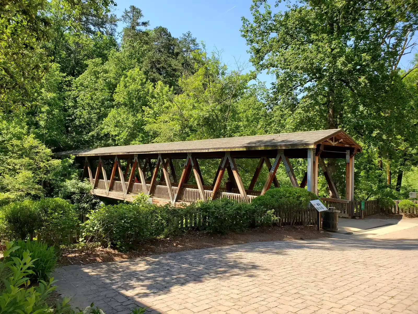 Side view of a long timber covered footbridge with a grey shingle roof and a row of reddish diagonal V-shaped trusses, its near end supported on a galvanised steel post, set in front of a tall wall of bright green deciduous trees and pines under a blue sky with a faint contrail. Below it are hedges and a picket fence, with a cobbled brick-paver plaza in the foreground, a small interpretive panel and a trash can at the entrance. One person, partly hidden, is on the bridge.