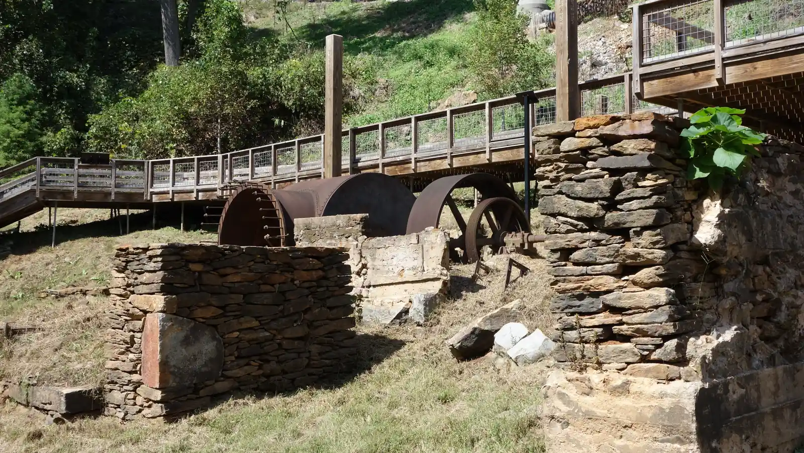 Ruins in a grassy clearing: low dry-stacked stone walls and a crumbling stone pier frame rusted industrial machinery, including a large riveted iron drum or wheel housing, a big flywheel and a spoked pulley wheel on a shaft. Behind and above, a wooden boardwalk with wire-mesh railings and tall timber posts zig-zags up a green, sunlit hillside. One dark interpretive sign post stands at centre right.