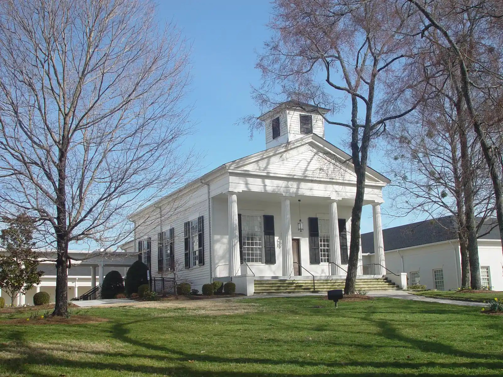 A white clapboard Greek Revival church with a four-column front portico, a triangular pediment and a square white belfry with louvred openings, black-shuttered tall windows, on a green lawn shaded by tall bare winter trees. Lower white modern wings extend to the left and right. Blue sky.