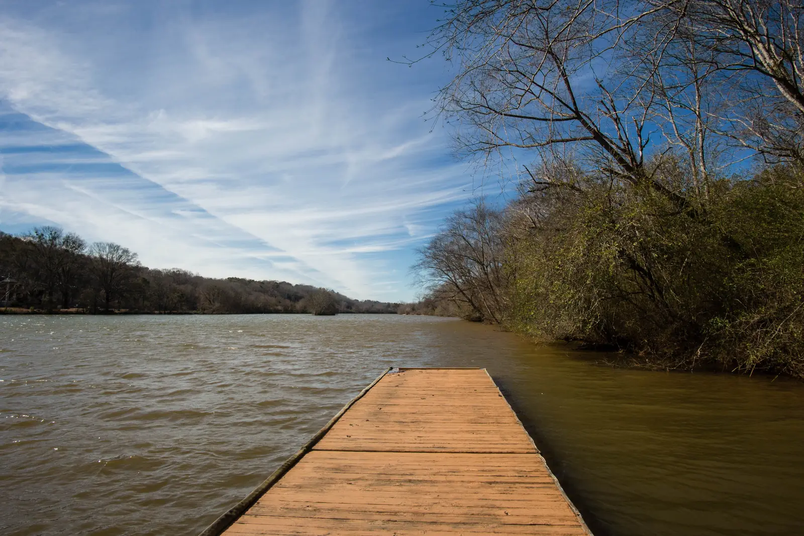 Low wide-angle view along a weathered orange-brown floating wooden dock that leads out into a wide, wind-rippled muddy-brown river. Bare winter trees and green-brown shrubs overhang the right bank, and a low wooded far shore runs to a vanishing point. Streaks of cirrus cloud and contrails radiate across a deep-blue sky.