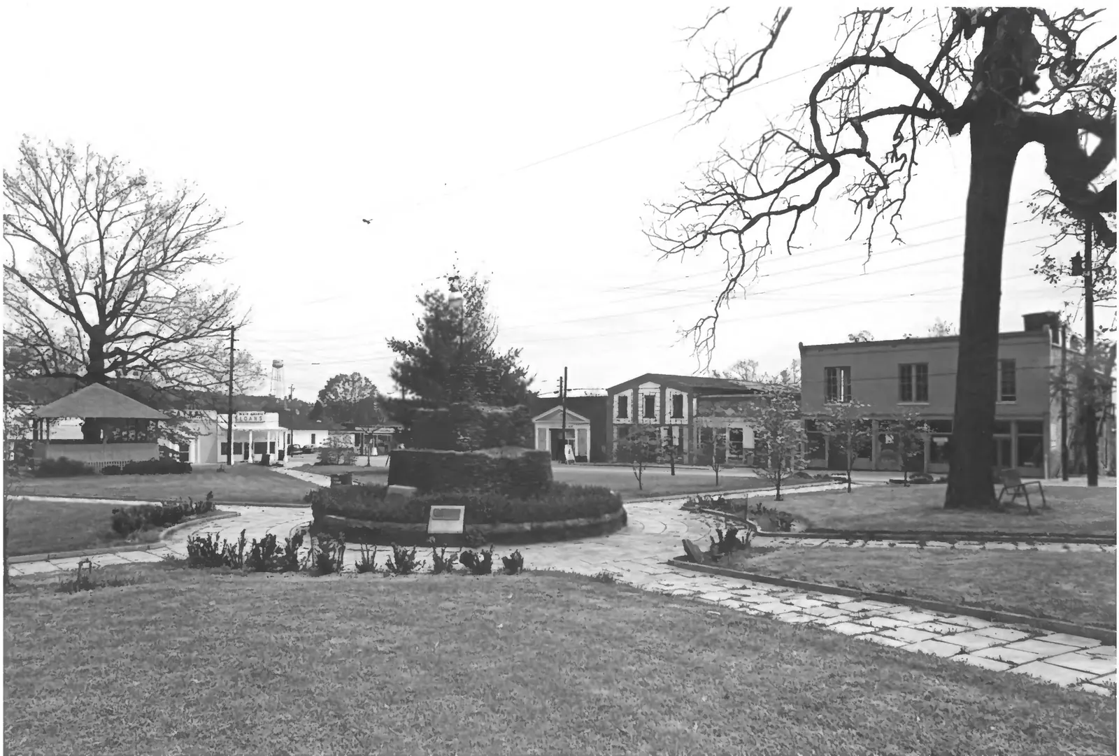 A black-and-white photograph across a grassy square: curving paved paths lead to a central round planting bed with a low monument and plaque, and a fir tree with a slender pole behind it. On the far side of the street is a row of old commercial buildings: a two-storey flat-roofed brick block at right, a stuccoed front with a stepped parapet, a small classical temple-front building, and at far left a white building with a "LOANS" sign. A water tower rises on the horizon, an open-sided bandstand-like shelter stands at left, and bare and leafing trees frame the scene.