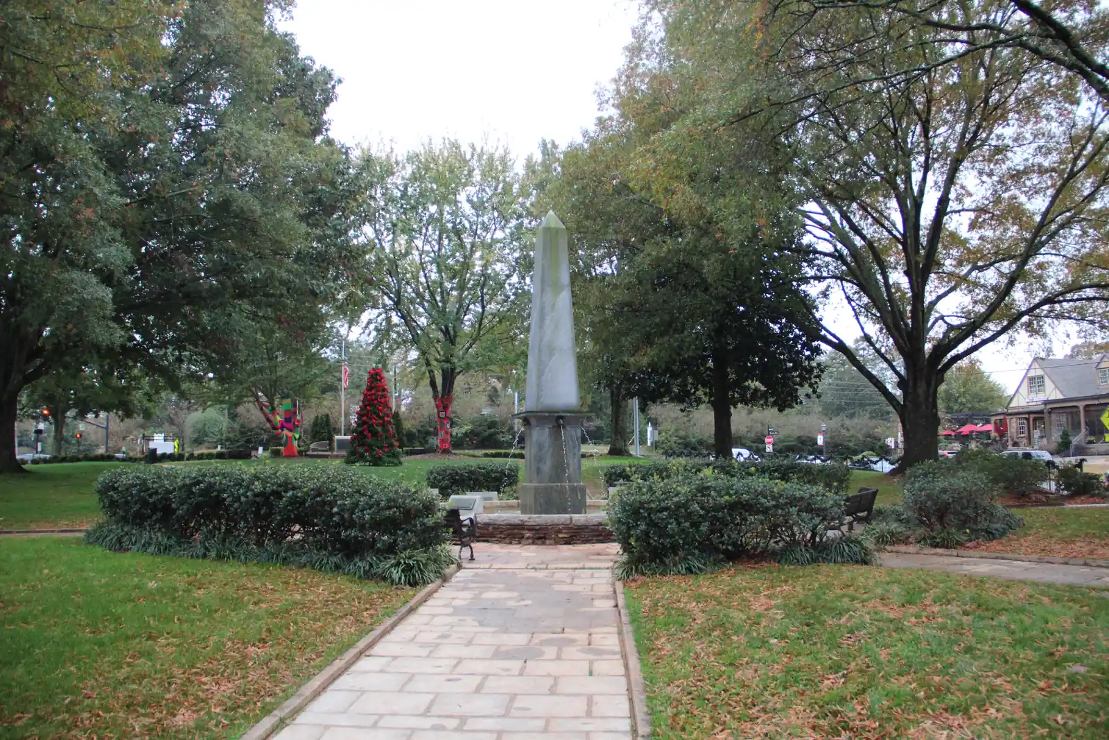 A stone-paved path leads between low clipped hedges to a tall granite obelisk fountain with water spouting from its base, in a leafy park ringed by large oaks in early autumn colour. On the left, trees are wrapped in bright knitted yarn and a red-ribboned holiday tree stands near a flagpole with an American flag. On the right edge, a cream half-timbered Tudor-style building with red patio umbrellas and parked cars is visible beyond a black iron fence. Overcast sky.