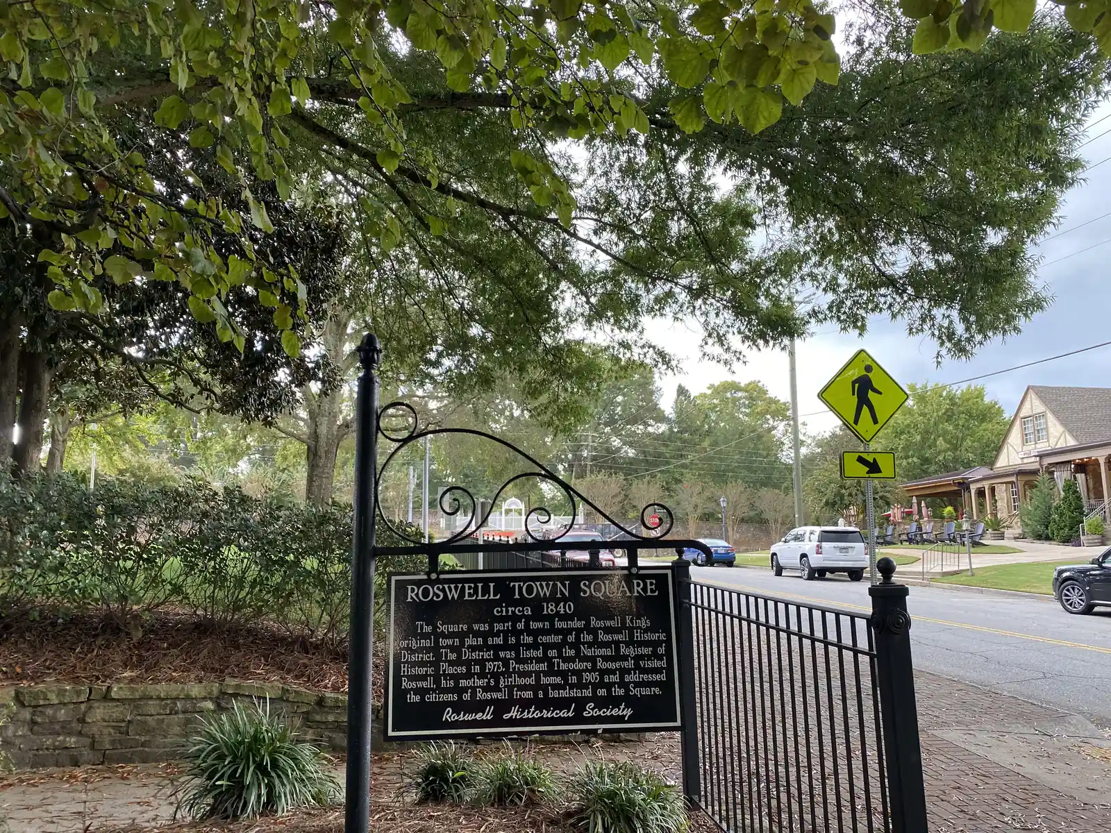 A black wrought-iron hanging sign beside an iron fence, under a leafy tree canopy, reads: "Roswell Town Square, circa 1840. The Square was part of town founder Roswell King's original town plan and is the center of the Roswell Historic District. The District was listed on the National Register of Historic Places in 1973. President Theodore Roosevelt visited Roswell, his mother's girlhood home, in 1905 and addressed the citizens of Roswell from a bandstand on the Square. Roswell Historical Society". Beyond it, a yellow pedestrian-crossing sign, a street with parked SUVs, and the porch of a stone-and-stucco Tudor-style building with Adirondack chairs.