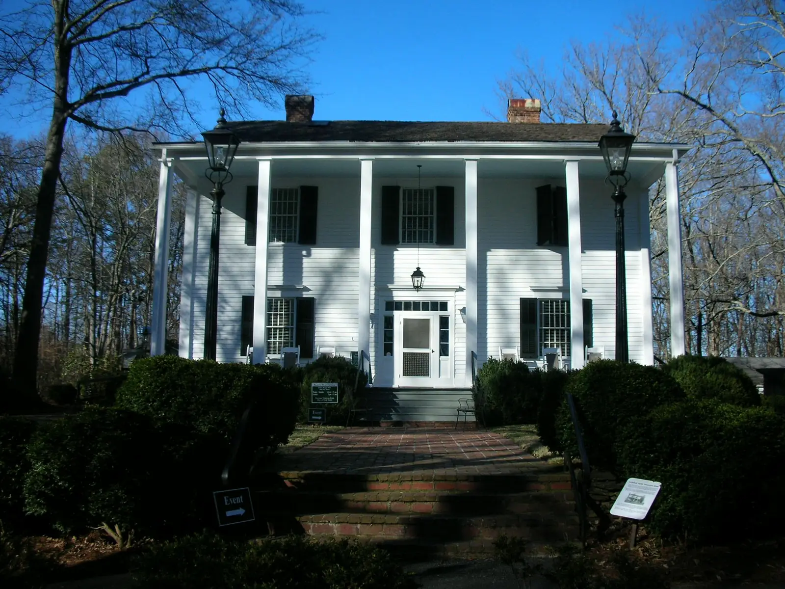 Straight-on winter view of a white two-storey clapboard house with a full-width two-storey porch of square columns, black shutters, two brick chimneys and a hanging lantern over a front door with a sidelit transom. Tall black gas-style lamp posts stand at each side of brick entry steps that rise through dark clipped boxwoods. Small signs read "Event" with arrows, a hours sign stands on the walk, and an interpretive panel with a historic photo is at lower right. Bare trees fill a deep blue sky.