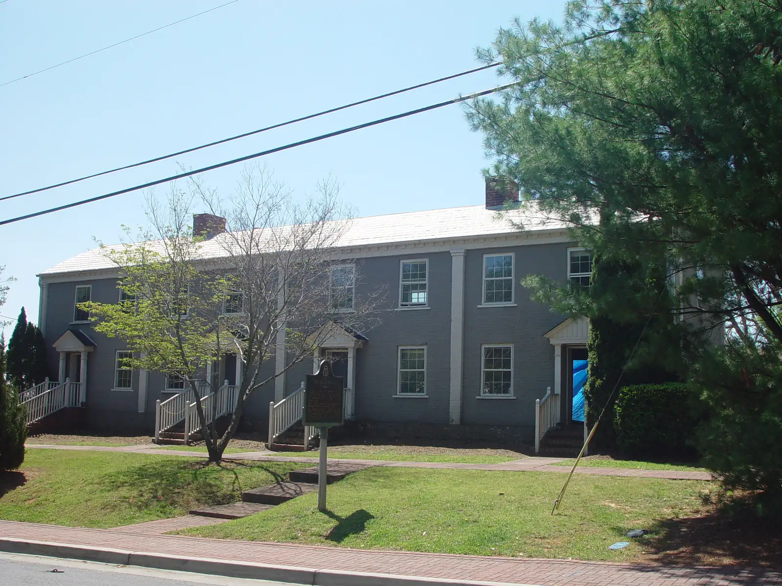 A long two-storey brick row building painted grey with white trim, white pedimented door hoods, white stair railings and two brick chimneys on a pale roof, set behind a sloping lawn and a brick sidewalk. A green-and-gold Georgia historical marker in front reads "The Old Bricks". A young bare-branched tree is on the left, a large pine overhangs the right, a blue tarp hangs in one doorway, and overhead power lines cross a clear blue sky.