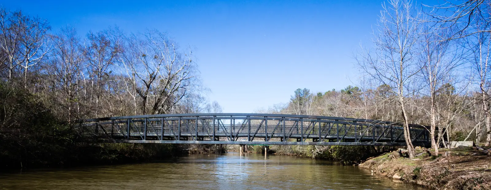 Wide panoramic view of a dark steel truss footbridge arching low across a calm brown creek, framed by tall bare winter trees and pale sycamores on both banks under a vivid cloudless blue sky. A rocky, leaf-covered bank is in the right foreground.